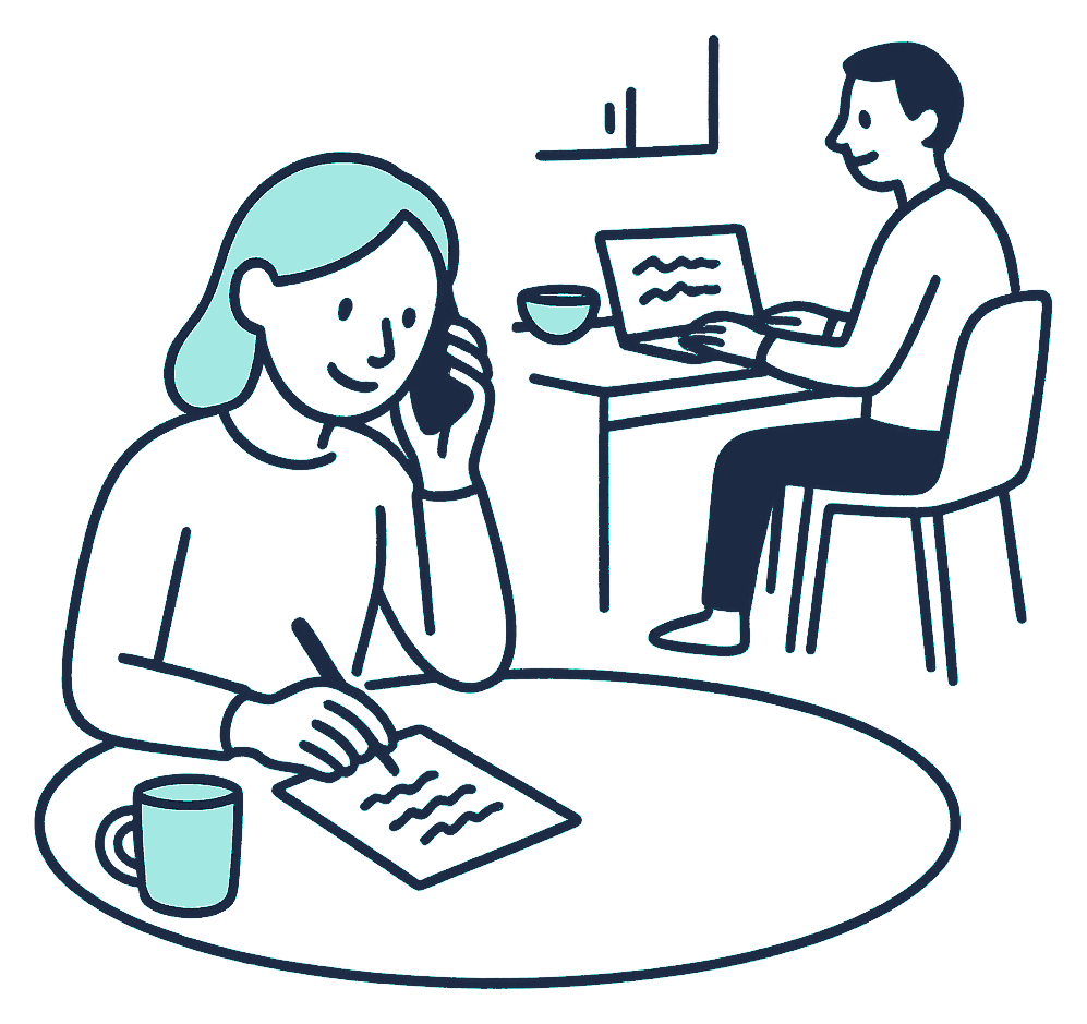 A person takes notes during a phone call at a kitchen table while, across the room, a second person fills in an application on a laptop