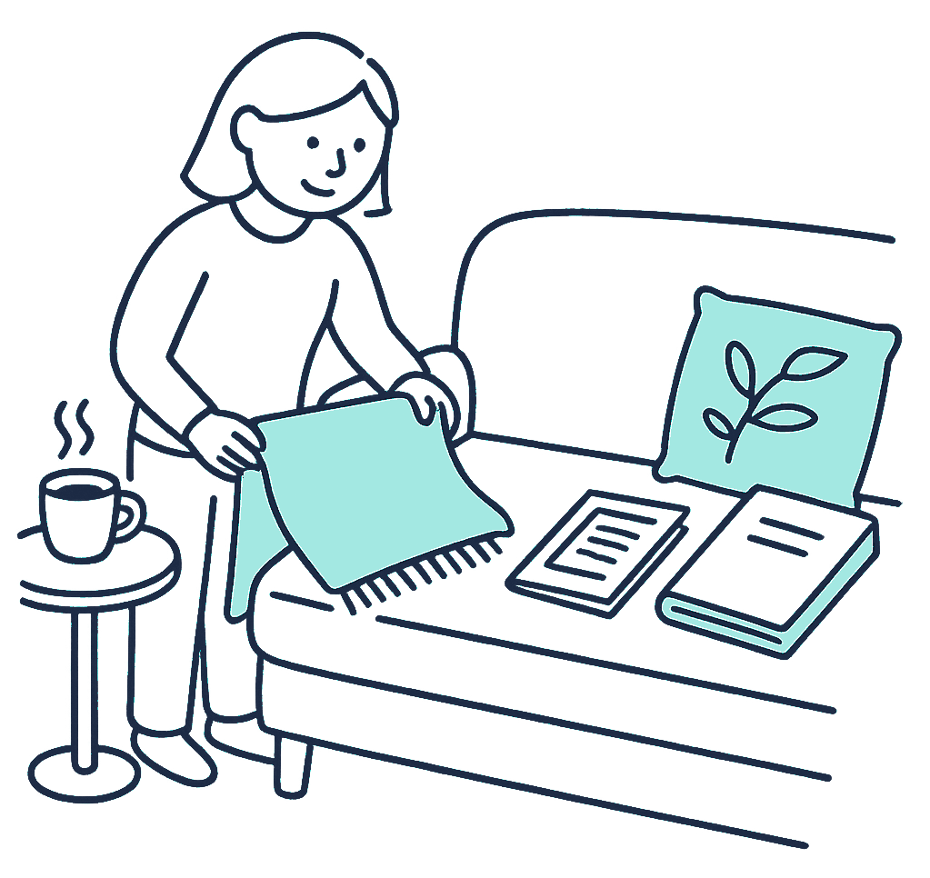 A person folds a large woollen blanket down to a smaller square on a couch beside a slim funeral-cover brochure and a thicker life insurance policy folder, a cup of tea on the side table