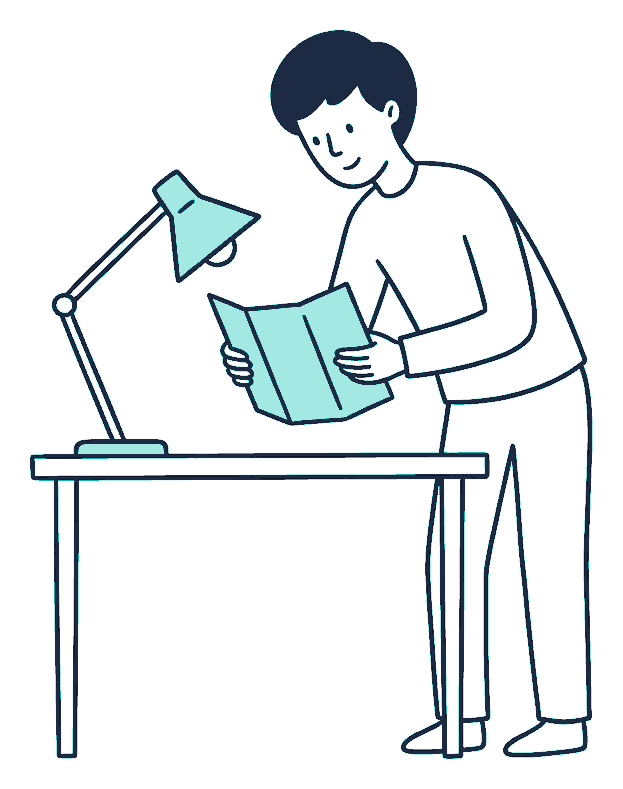 A person leans over a desk reading a plain folded pamphlet under an angled desk lamp