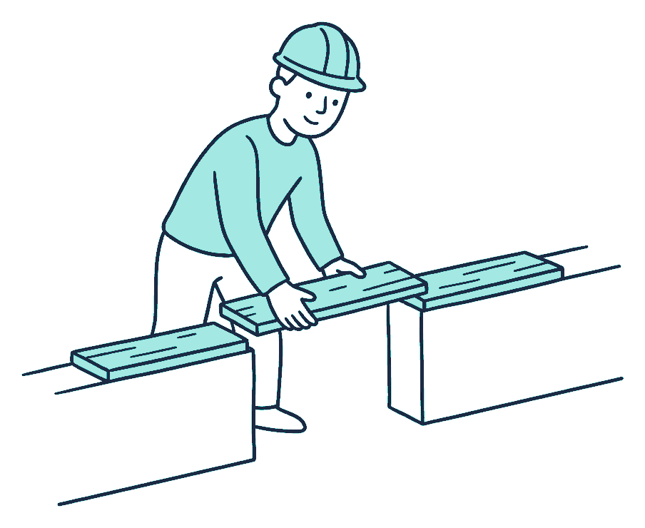 A builder lays three short planks end to end across a gap between two low walls